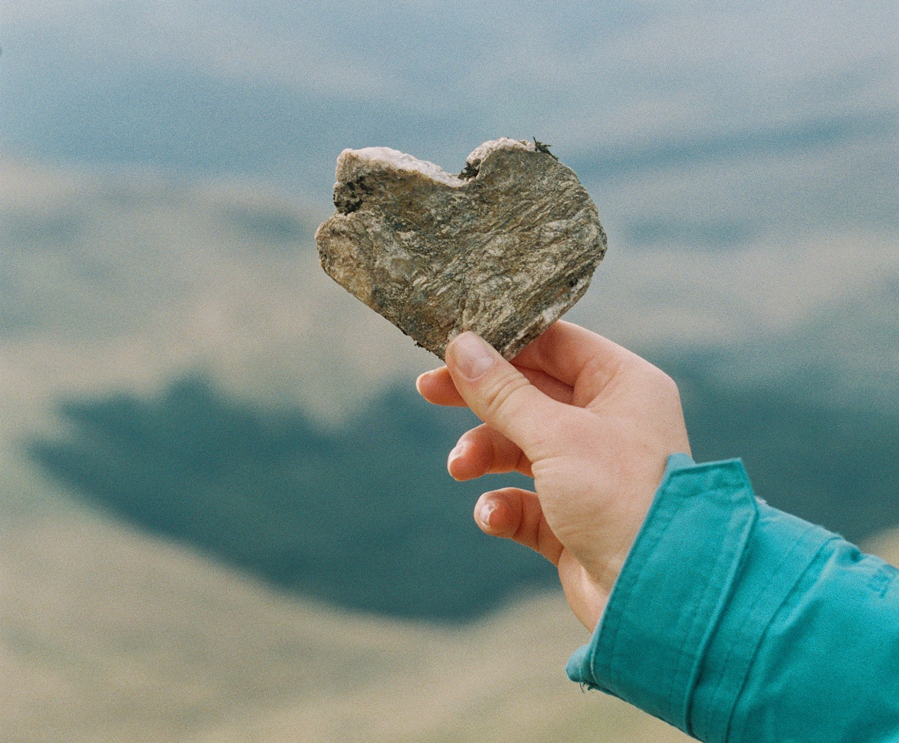A hand holding a heart-shaped rock in front a distant landscape